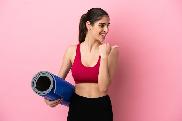 Joven mujer caucásica de deporte que va a clases de yoga mientras sostiene una estera apuntando hacia el lado para presentar un producto