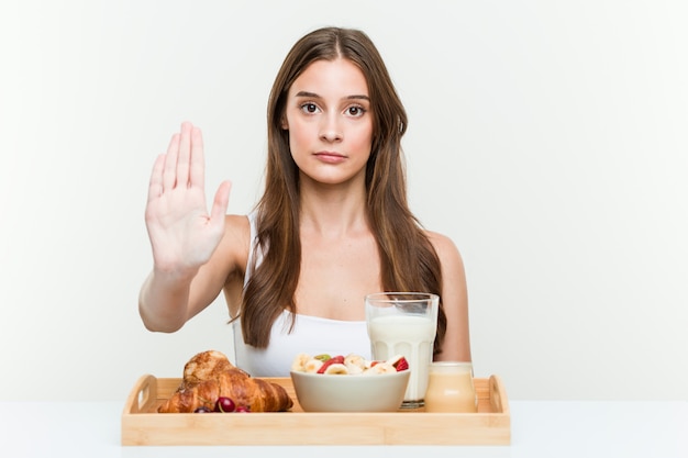 Joven mujer caucásica desayunando de pie con la mano extendida que muestra la señal de stop