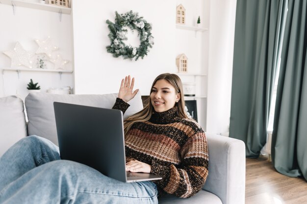 Joven mujer caucásica desea feliz navidad por videollamada usando computadora portátil en vacaciones en casa en el sofá.