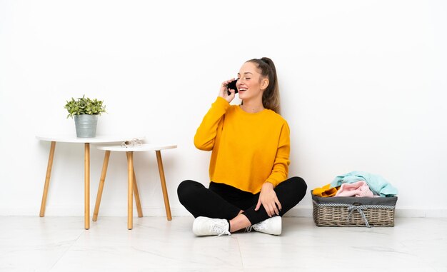 Joven mujer caucásica doblando ropa sentada en el suelo aislada de fondo blanco manteniendo una conversación con el teléfono móvil