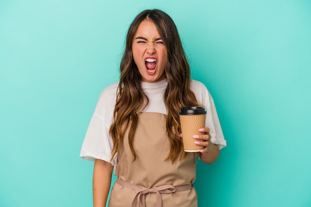 Joven mujer caucásica empleado de tienda sosteniendo un café para llevar aislado sobre fondo azul gritando muy enojado y agresivo.