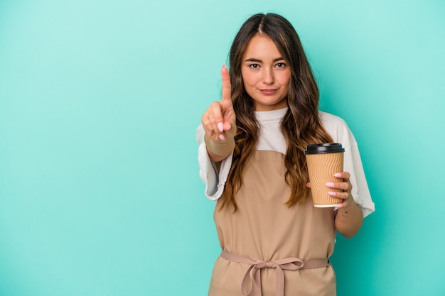 Joven mujer caucásica empleado de tienda sosteniendo un café para llevar aislado sobre fondo azul mostrando el número uno con el dedo.