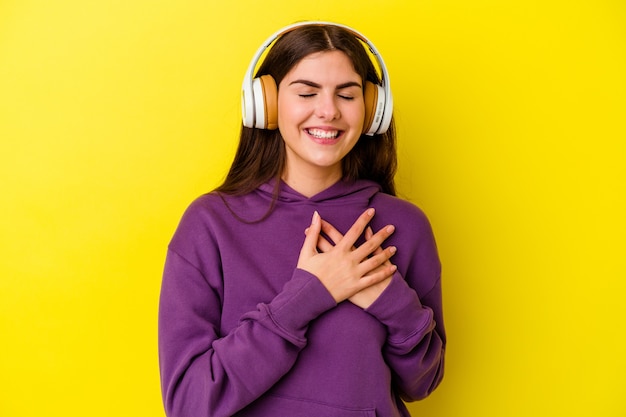 Joven mujer caucásica escuchando música con auriculares en rosa riendo manteniendo las manos en el corazón, concepto de felicidad.