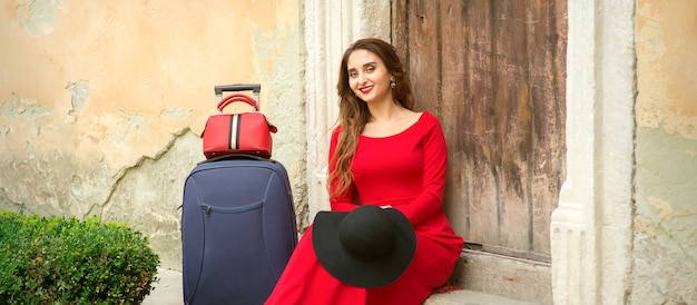 Una joven mujer caucásica está sentada en el umbral de una casa antigua con un vestido largo rojo con una maleta