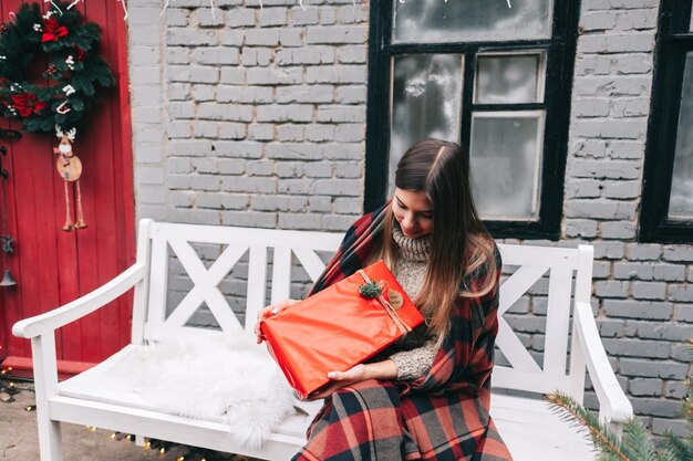 Joven mujer caucásica está sosteniendo un regalo de Navidad mientras está sentado en un banco en el patio trasero.