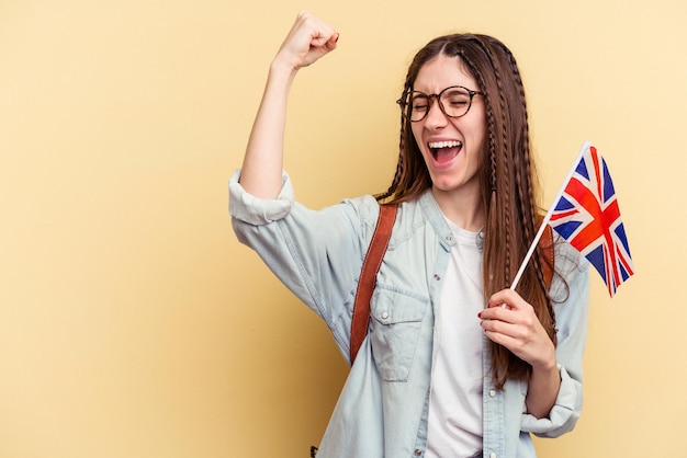 Joven mujer caucásica estudiando inglés aislada de fondo amarillo levantando el puño después de un concepto ganador de la victoria