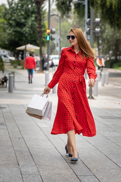 Joven mujer caucásica feliz en vestido rojo caminando con estilo por la calle llevando bolsas de la compra.