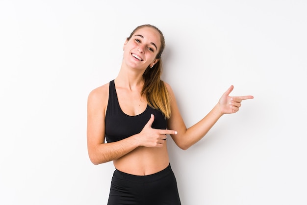 Joven mujer caucásica fitness posando en un blanco emocionado apuntando con los dedos índices de distancia.
