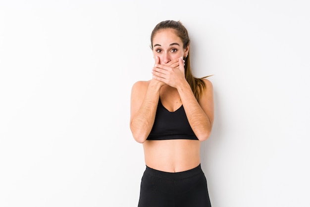 Joven mujer caucásica fitness posando en una pared blanca conmocionado cubriendo la boca con las manos.