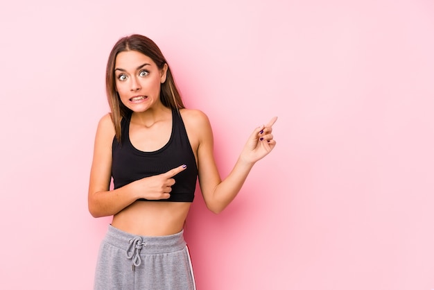Joven mujer caucásica fitness posando sobre fondo rosa sorprendido apuntando con los dedos índices a un espacio de copia.