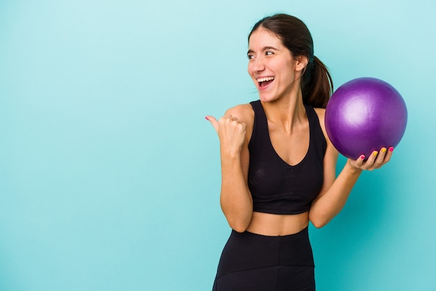 Joven mujer caucásica fitness sosteniendo una bola aislada sobre fondo azul apunta con el dedo pulgar lejos, riendo y sin preocupaciones.