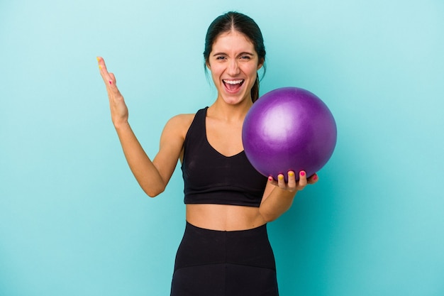 Foto joven mujer caucásica fitness sosteniendo una bola aislada sobre fondo azul recibiendo una agradable sorpresa, emocionada y levantando las manos.