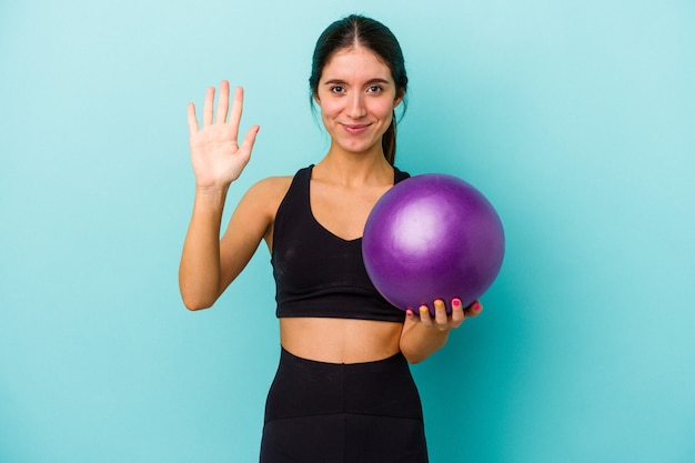 Joven mujer caucásica fitness sosteniendo una bola aislada sobre fondo azul sonriendo alegre mostrando el número cinco con los dedos.