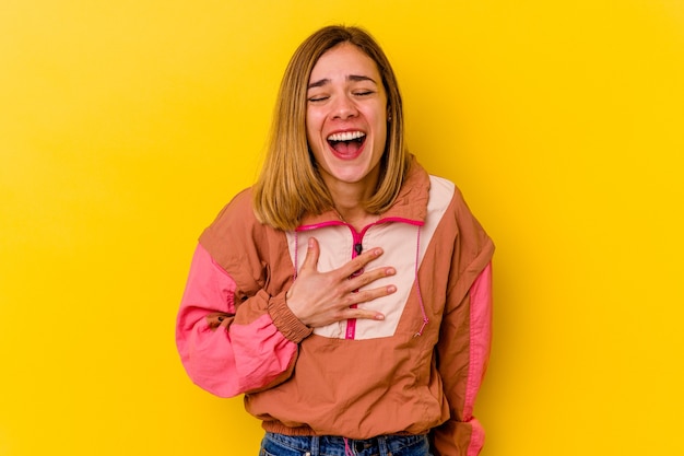 Joven mujer caucásica flaca aislada en amarillo se ríe a carcajadas manteniendo la mano en el pecho.