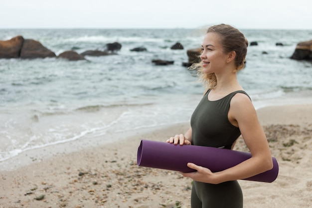 Joven mujer caucásica en forma con colchoneta de yoga en la playa Deportes al aire libre