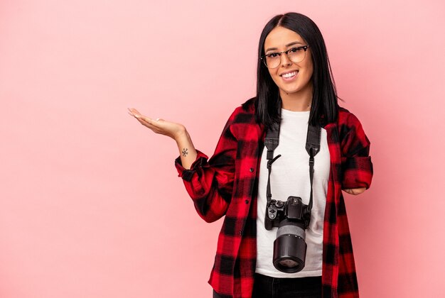 Joven mujer caucásica de fotografía con un brazo aislado sobre fondo rosa mostrando un espacio de copia en una palma y sosteniendo otra mano en la cintura.