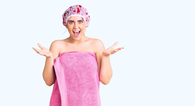 Joven mujer caucásica con gorro de ducha y toalla celebrando la victoria con una sonrisa feliz y una expresión ganadora con las manos levantadas