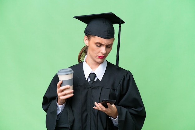 Joven mujer caucásica graduada universitaria sobre antecedentes aislados sosteniendo café para llevar y un móvil