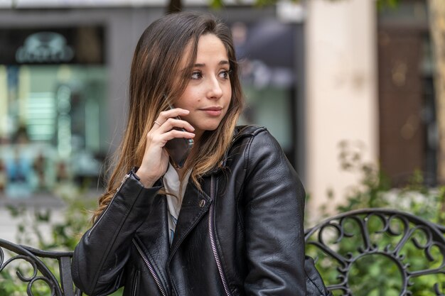 Joven mujer caucásica hablando felizmente con su teléfono inteligente sentado en un banco