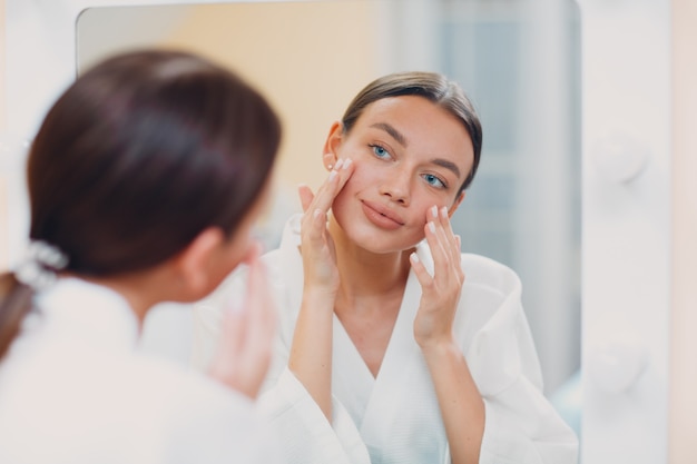 Joven mujer caucásica haciendo gimnasia facial de yoga de construcción facial masaje de estiramiento facial linfático en casa espejo