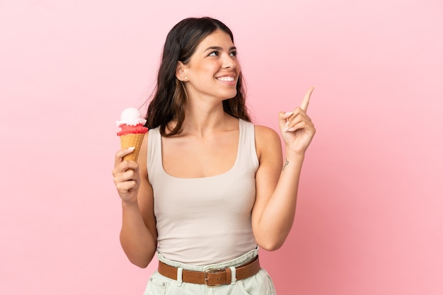 Joven mujer caucásica con un helado de cucurucho aislado sobre fondo rosa apuntando hacia una gran idea