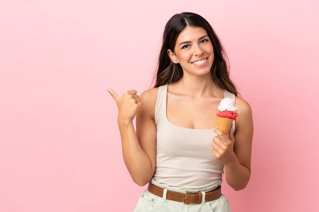 Joven mujer caucásica con un helado de cucurucho aislado sobre fondo rosa apuntando hacia el lado para presentar un producto