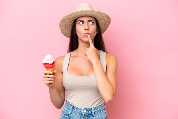 Joven mujer caucásica con un helado de cucurucho aislado sobre fondo rosa con dudas mientras mira hacia arriba