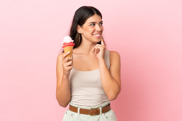 Joven mujer caucásica con un helado de cucurucho aislado sobre fondo rosa pensando en una idea mientras mira hacia arriba