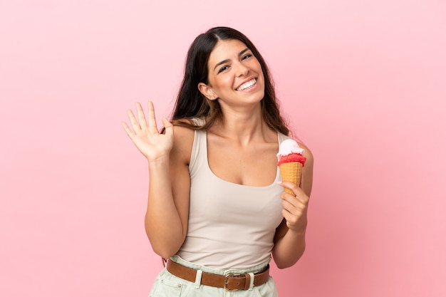 Joven mujer caucásica con un helado de cucurucho aislado sobre fondo rosa saludando con la mano con expresión feliz