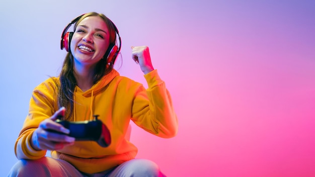 Una joven mujer caucásica hermosa con una capucha amarilla jugando un videojuego usando un joystick y auriculares con una divertida sorpresa de enfoque y una cara de victoria ganadora con luz de neón azul y roja