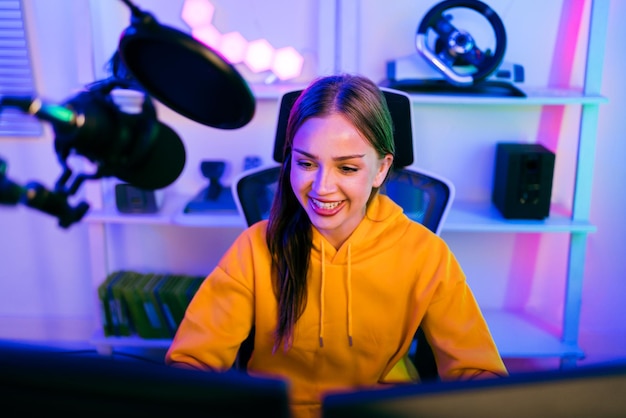 Joven mujer caucásica jugador profesional con capucha amarilla se sienta en una silla con una mesa de juegos con micrófono de monitor de teclado de PC Prepárate para la competencia juego de elenco o grabar un podcast