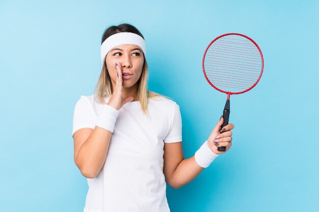 Joven mujer caucásica jugando bádminton aislado está diciendo una noticia secreta de frenado en caliente y mirando a un lado