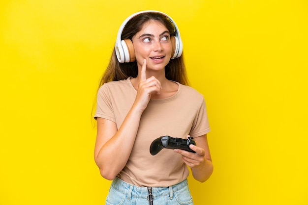 Joven mujer caucásica jugando con un controlador de videojuegos aislado en un fondo amarillo pensando en una idea mientras mira hacia arriba