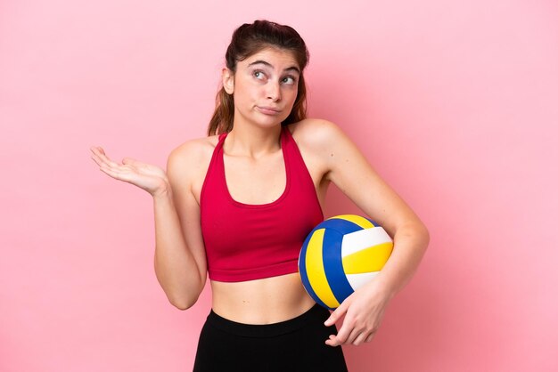 Joven mujer caucásica jugando voleibol aislada de fondo rosa que tiene dudas mientras levanta las manos