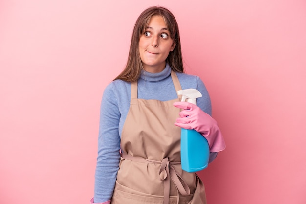 Joven mujer caucásica limpiador sosteniendo spray aislado sobre fondo rosa confundido, se siente dudoso e inseguro.