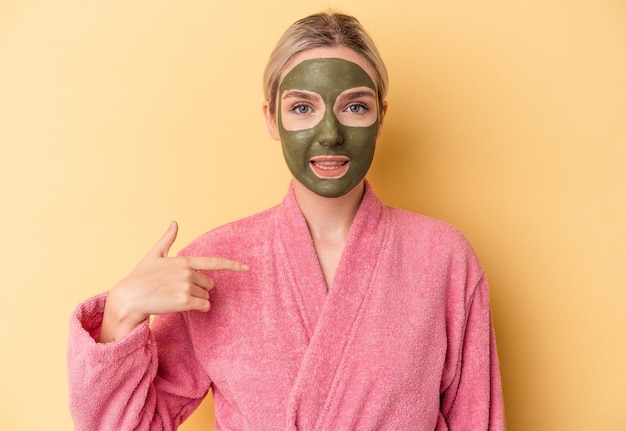 Joven mujer caucásica con máscara facial aislada sobre fondo amarillo persona apuntando con la mano a un espacio de copia de camisa, orgulloso y seguro