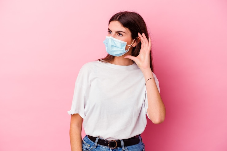 Foto joven mujer caucásica con una máscara para virus aislado en pared rosa tratando de escuchar un chisme.