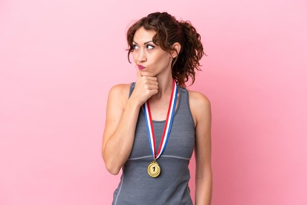 Joven mujer caucásica con medallas aisladas en un fondo rosado que tiene dudas y con una expresión facial confusa