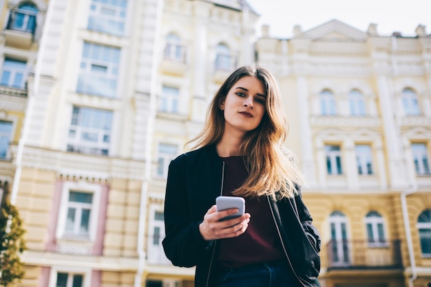 joven mujer caucásica mirando a la cámara y posando mientras espera a un amigo en la calle con teléfono móvil