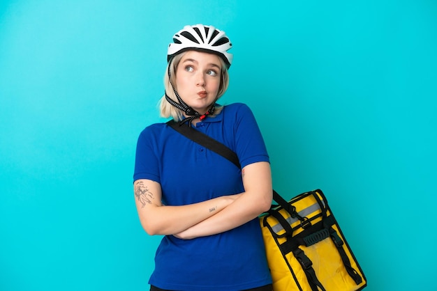 Joven mujer caucásica con mochila térmica aislada de fondo azul haciendo gestos de duda mientras levanta los hombros