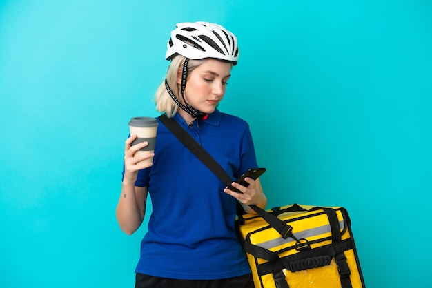 Joven mujer caucásica con mochila térmica aislado sobre fondo azul con café para llevar y un móvil