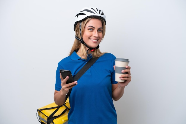 Joven mujer caucásica con mochila térmica aislado sobre fondo blanco sosteniendo café para llevar y un móvil