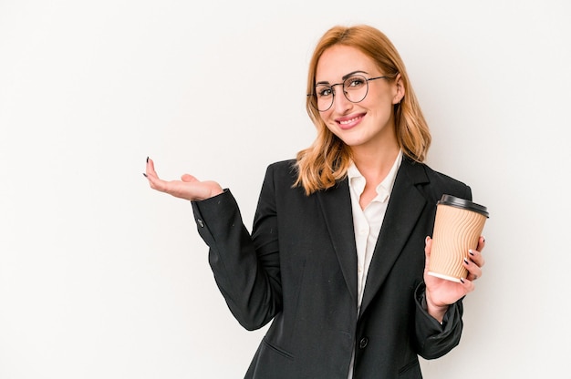 Joven mujer caucásica de negocios sosteniendo café para llevar aislado en fondo blanco mostrando un espacio de copia en una palma y sosteniendo otra mano en la cintura.