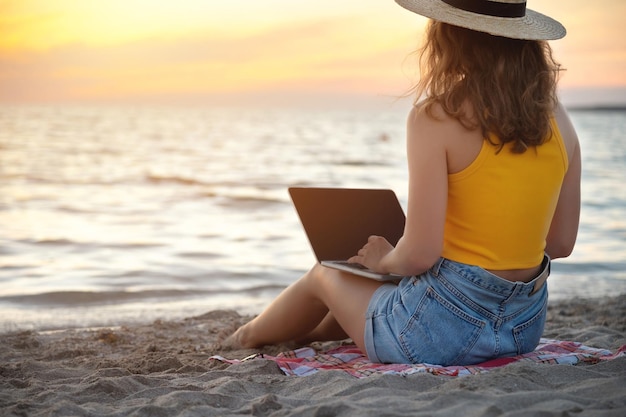 Joven mujer caucásica en pantalones cortos y sombrero en la playa sentada en la arena navegando por las redes sociales