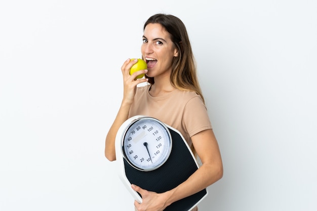 Joven mujer caucásica en pared blanca con máquina de pesaje y con una manzana