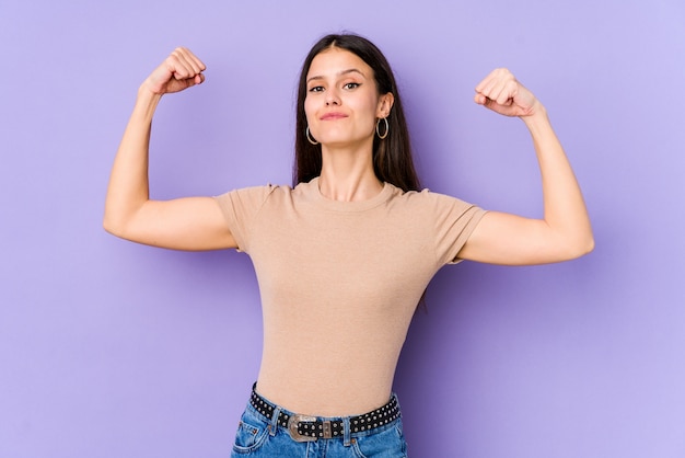 Joven mujer caucásica en pared púrpura que muestra gesto de fuerza con los brazos, símbolo del poder femenino