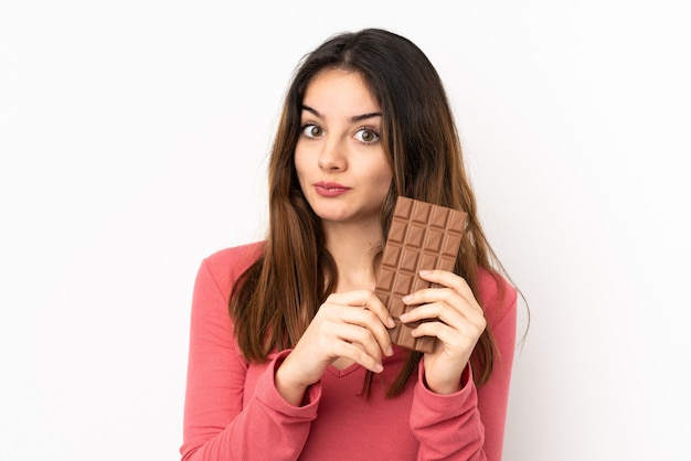 Joven mujer caucásica en pared rosa tomando una tableta de chocolate y teniendo dudas