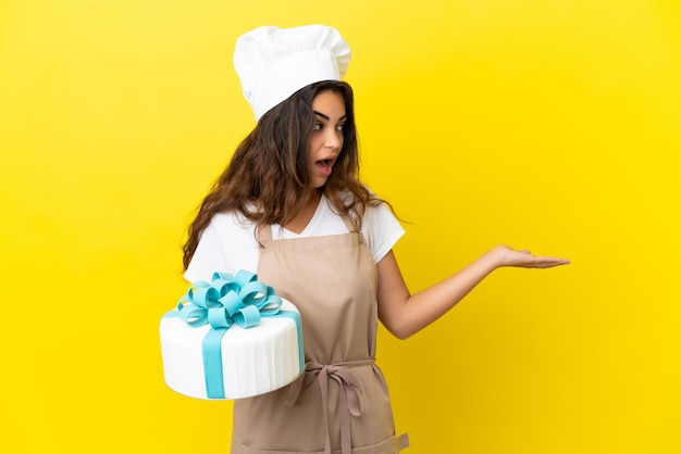 Joven mujer caucásica pastelera con un gran pastel aislado sobre fondo amarillo con expresión de sorpresa mientras mira de lado