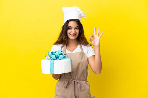 Joven mujer caucásica pastelera con un gran pastel aislado sobre fondo amarillo que muestra el signo de ok con los dedos