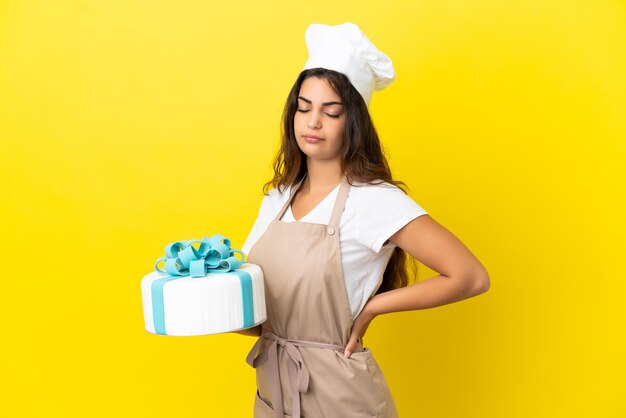 Joven mujer caucásica pastelera con un gran pastel aislado sobre fondo amarillo que sufre de dolor de espalda por haber hecho un esfuerzo